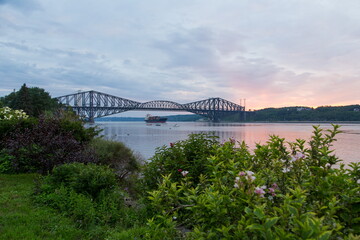 
Sunset view of small boats and large merchant vessel under and near the Quebec Bridge over the St. Lawrence River seen from a south shore garden in soft focus foreground, Lévis, Quebec, Canada
