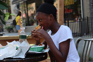Girl eating hamburger sandwich while sitting outdoors while sitting  at table on city sidewalk