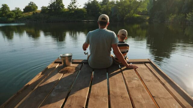 Rear view of a father with his little son are fishing while sitting on a wooden pier by the pond