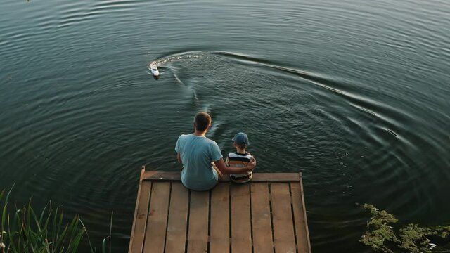 Top view from the back of a dad with a little son sitting on a wooden pier playing with a RC speed-boat on the lake