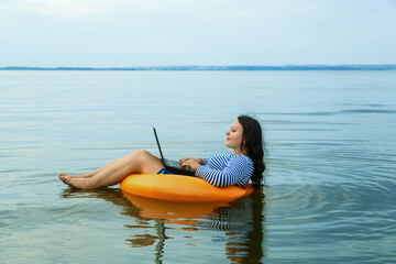 Woman on vacation works remotely with a laptop in a swimming circle in the morning water.
