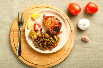 A plate with vegetables and a piece of chicken baked on a round stand on a wooden table with a fork near the garlic and tomatoes.