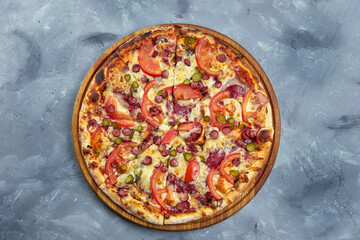 Baked italian pizza with smoked sausages, pickled cucumbers, salami and tomatoes on a wooden tray on a gray background.