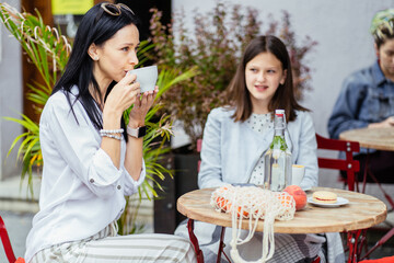 Mom and her teenager grown up daughter drink coffee, stiitng at outdoor cafe, trying socializing. separately from elderly mother, parent think about bad relations and misunderstandings with child.