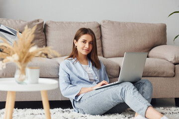 Beautiful young woman in jeans and blue shirt sits on the floor with laptop indoors at home