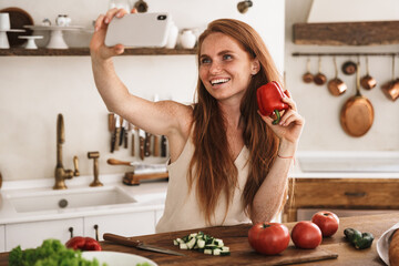 Image of happy woman taking selfie photo on mobile phone while cooking