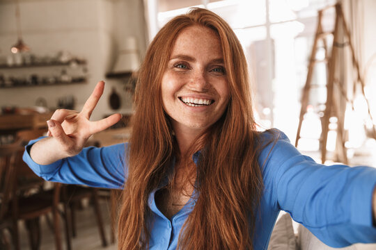 Image of happy ginger woman showing peace sign and taking selfie photo