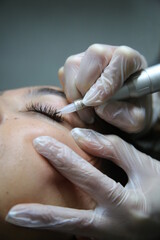 woman lying on couch at beauty salon with closed eyes and enjoying when cosmetologist making eyeliner and  eyebrow  permanent make up. Professional wearing black gloves and holding speciality tool.