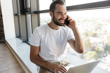 Image of young joyful man using laptop and talking on cellphone
