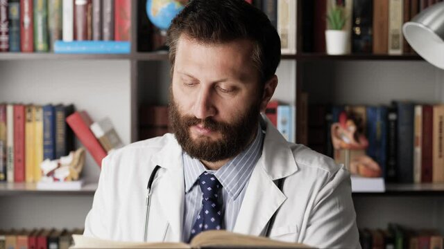 Doctor is reading book. Male doctor in white coat at workplace in hospital office sits at table and reads large paper book, turns page
