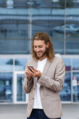 Portrait of handsome curly businessman in casual wear holding smartphone and smiling.Successful manager using mobile phone apps,texting message,browsing internet,looking at phone near business center.