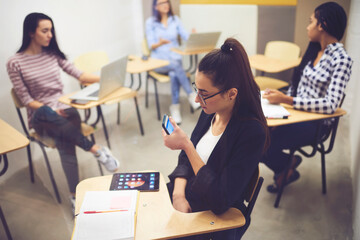 Concentrated young female student sitting in classroom with members of group preparing for lesson while using smartphone for browsing information on internet,young team sitting at desks doing homework