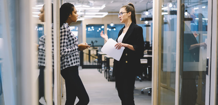 Two female colleagues having friendly conversation during informal meeting in office discussing working process, project manager of media marketing company giving advice to young afro american trainee