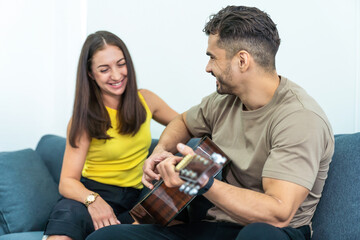 Young couple sitting smiling, playing guitar together on the sofa at home. Concept of the family spend time together, happy, quarantined 