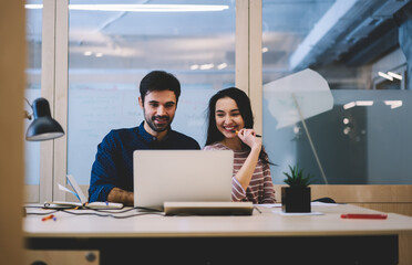 Positive bearded male and cheerful attractive female dressed in casual outfit having fun while reading news on laptop connected to wifi.Smiling good looking colleagues working together on gadget