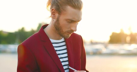 Caucasian young handsome man in red jacket writing his plans in notebook. Attractive male noting in planner at car parking. Leaving notes and planning. Cars on background.