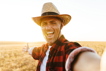 Fototapeta premium Image of excited adult man in straw hat taking selfie photo while standing at cereal field