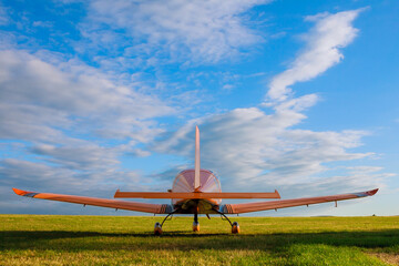 A small 2-seater plane against a blue sky with white clouds. Back view.