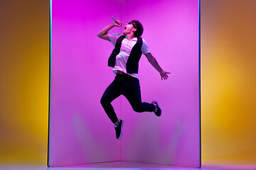 Fototapeta na wymiar In high jump. Young male musician, singer performing on pink-orange background in neon light. Concept of music, hobby, festival, entertainment, emotions. Joyful party host, singer, portrait of artist.