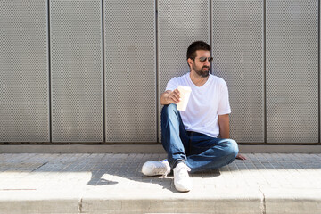 Bearded man with sunglasses sitting on ground while holding a take away coffee