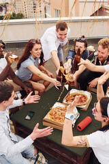 Doing cheers by bottles with beer. Group of young people in casual clothes have a party at rooftop...