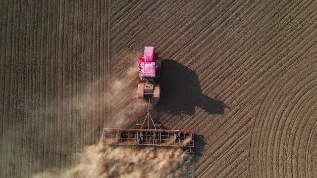 Shooting from drone flying over tractor with harrow system plowing ground on cultivated farm field, pillar of dust trails behind, preparing soil for planting new crop, agriculture concept, top view.