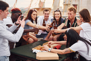 Doing cheers by bottles with beer. Group of young people in casual clothes have a party at rooftop together at daytime