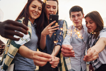 With sparklers in hands. Group of young people in casual clothes have a party at rooftop together at daytime