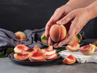 Women's hands hold a whole peach above the table on which there are whole and cut peaches.