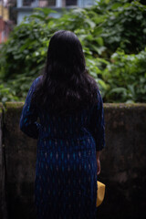 Backside view of an Indian woman standing in front of a green bush wearing a blue dress in a cloudy afternoon. Indian lifestyle