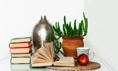 Naklejka na ściany i meble Cozy home interior decor: cup of coffee, stack of books, plants in pots, open book on a wicker stand, pillows on a white table. Distance home education.Quarantine concept of stay home