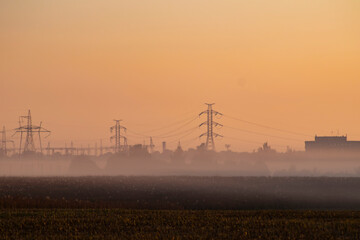 A beautiful misty industrial landscape with orange sunrise sky, white fog, distribution electric substation with power lines and transformers and trees and an off focus field in the foreground