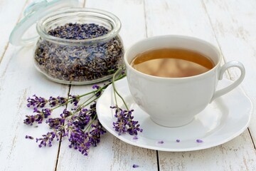 A Cup of tea with lavender flowers and a jar of dried lavender on a white wooden table close up