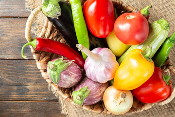 Various vegetables in a basket on a brown wooden table. Lots of raw vegetables in the basket. Eggplant, tomatoes, garlic, sweet peppers, onions on the table. Top view with space for text. Healthy food