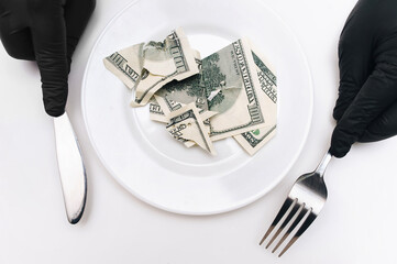 Hands in black gloves on a white tablecloth hold a fork and knife, between which stands a plate with torn dollars. The concept of a business lunch, fraud, scam and theft of capital.