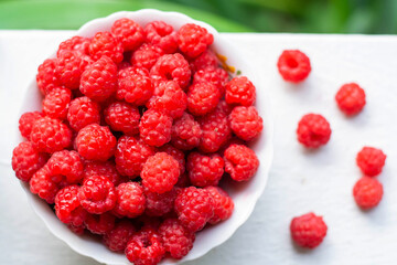 A white cup with fresh ripe raspberries stands against a background of bushes. Natural organic.