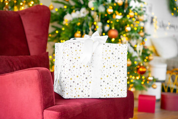 Beautiful gift box in white wrapping paper with stars, tied by silk ribbon with bow, stands on soft cozy armchair on holiday evening, decorated christmas tree on blurred background