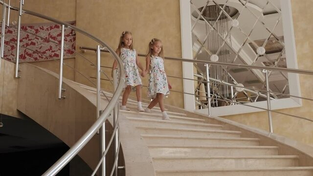 Two twin sisters in identical dresses descend the luxurious marble staircase in the hotel.