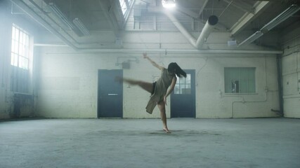 Beautiful dancer whips her hair, dancing passionately surrounded by mist in industrial warehouse