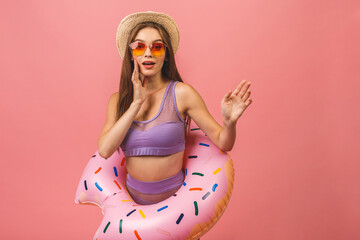 Portrait of a happy young woman dressed in swimsuit jumping and holding swim inflatable ring isolated over pink background.