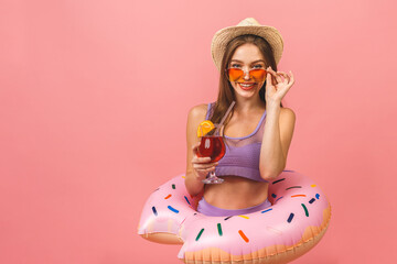 Portrait of a happy young woman dressed in swimsuit holding swim inflatable ring and cocktail drink isolated over pink background.