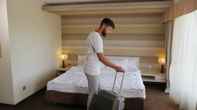 A tired bearded man enters the hotel rooms and falls on the bed. Exhausted young businessman lies with outstretched arms on bed next to suitcase. Entrepreneur tired after traveling, jet lag.