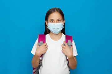 Brunette girl in white T-shirt in medical mask with backpack on blue background while studying at school during the pandemic 