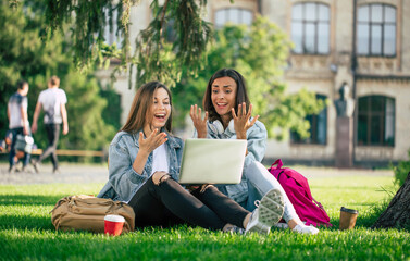 Two happy beautiful young student girl friends in casual denim clothes are relaxing in college park with laptop and smartphone on university background and drinking coffee.