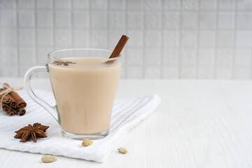 Hot indian masala chai or mixed spice tea made of milk and variety of aromatic herb such as anise and cinnamon in glass cup on towel on white wooden background at kitchen. Image with copy space