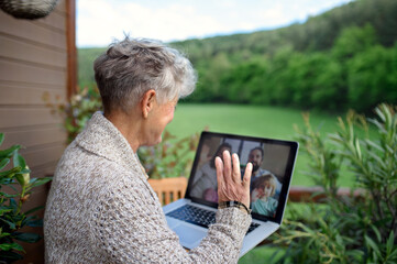 Senior woman with laptop sitting on terrace in summer, video call with family.
