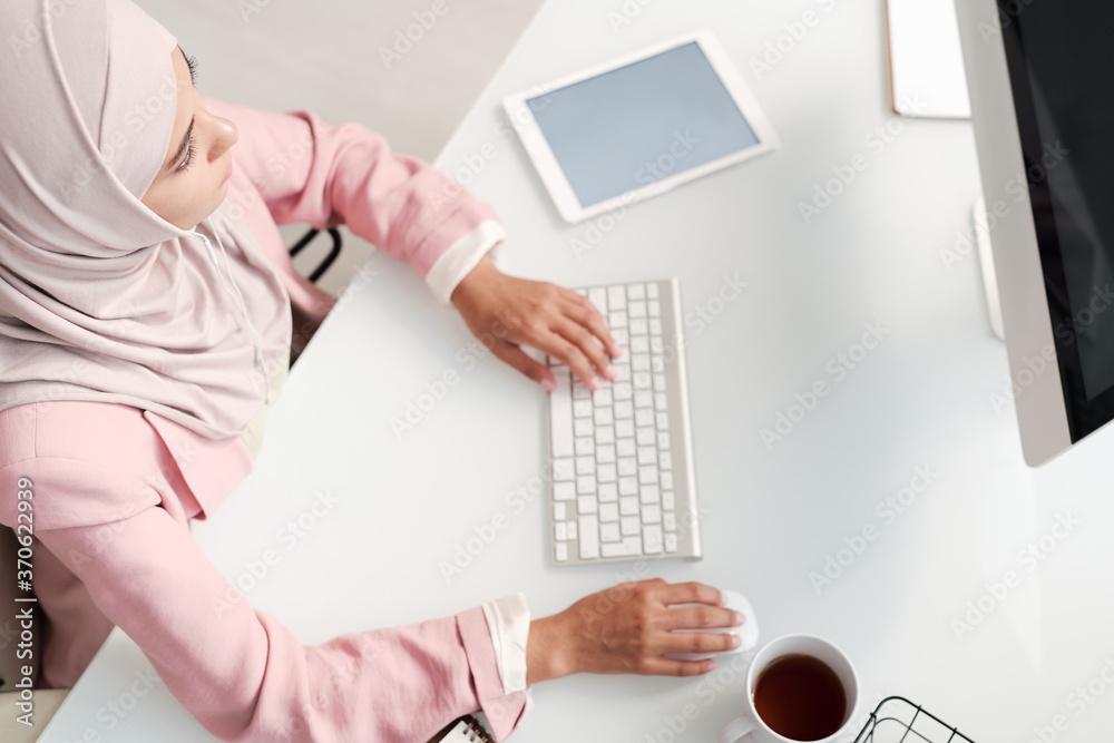 Poster Above view of busy young Muslim woman in hijab sitting at desk and working on project using computer