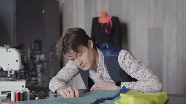Handsome brunette man yawning and putting head on the table next to sewing machine. Portrait of tired Caucasian designer or tailor overworking in atelier in the evening. Exhaustion and tiredness.
