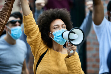 Black woman with protective face mask shouting through megaphone on public demonstrations.