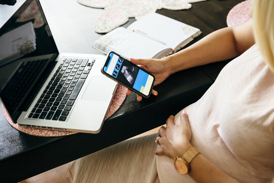 Pregnant black woman working in home office and looking at sonogram on phone